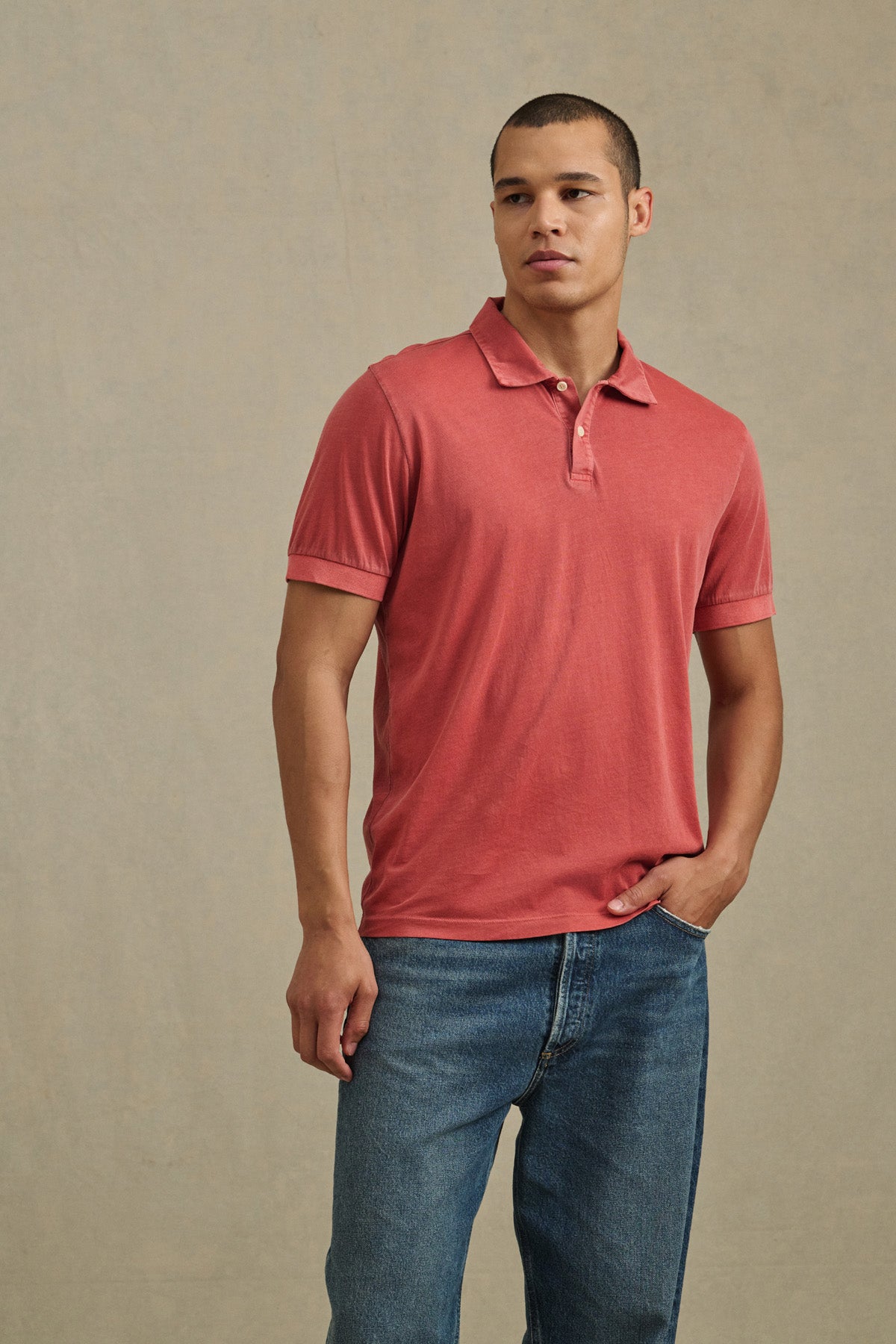 A person wears the ARROW POLO by Velvet by Graham & Spencer, a short-sleeved classic style in red, paired with blue jeans, standing with one hand in their pocket against a plain background.