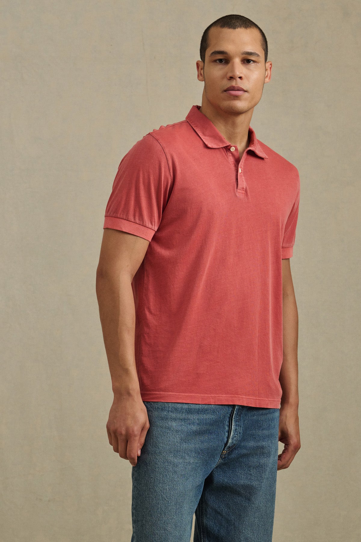 A man wears the ARROW POLO by Velvet by Graham & Spencer—a classic, short-sleeve red polo made from lightweight cotton—paired with blue jeans against a plain light background.