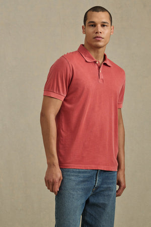 A man wears the ARROW POLO by Velvet by Graham & Spencer—a classic, short-sleeve red polo made from lightweight cotton—paired with blue jeans against a plain light background.