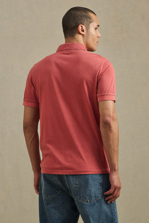 A man faces away, wearing the ARROW POLO by Velvet by Graham & Spencer in red with short sleeves and blue jeans, set against a plain beige background.