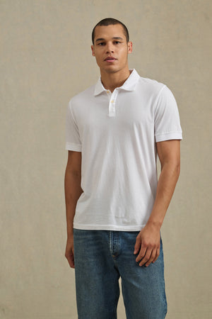 A man wearing the ARROW POLO by Velvet by Graham & Spencer and blue jeans stands against a neutral beige background.
