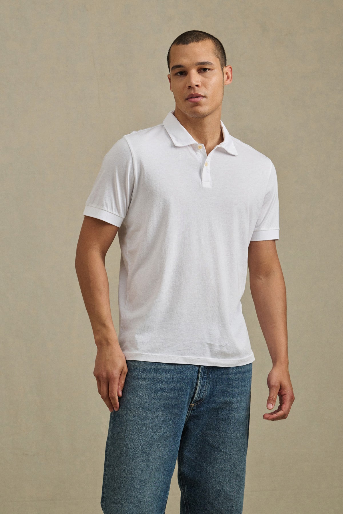 A man stands against a plain backdrop, facing the camera with a neutral expression, wearing the ARROW POLO by Velvet by Graham & Spencer in white and paired with blue jeans.