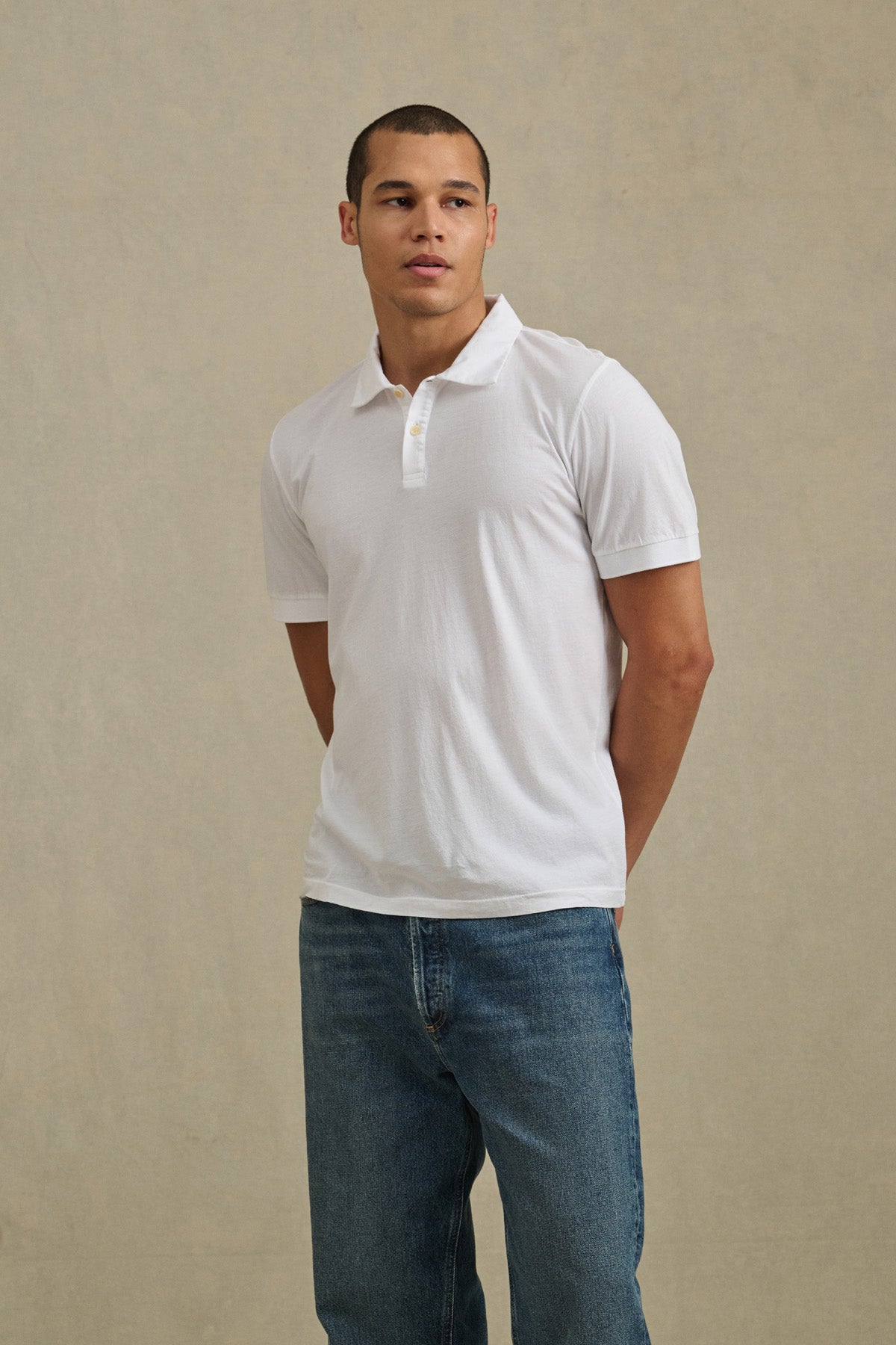 A man stands against a plain beige background with his hands behind his back, wearing the ARROW POLO by Velvet by Graham & Spencer in white and a pair of blue jeans.-41338467942593