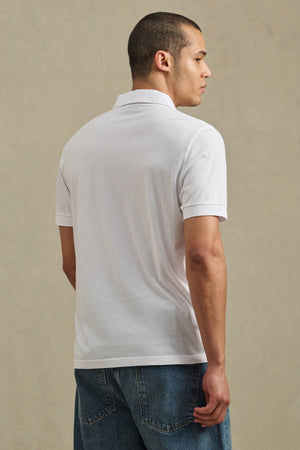 Someone wearing the ARROW POLO by Velvet by Graham & Spencer, a white cotton short-sleeve shirt, and blue jeans stands with their back to the camera against a neutral background.
