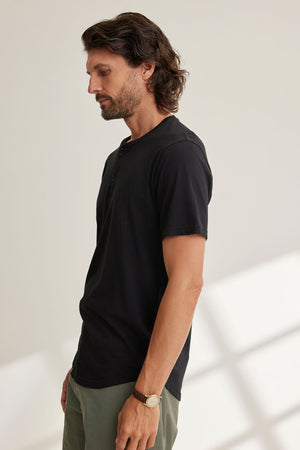 A long-haired man stands sideways, wearing a black FULTON HENLEY by Velvet by Graham & Spencer with green pants. He glances down, his watch catching the light on his left wrist.