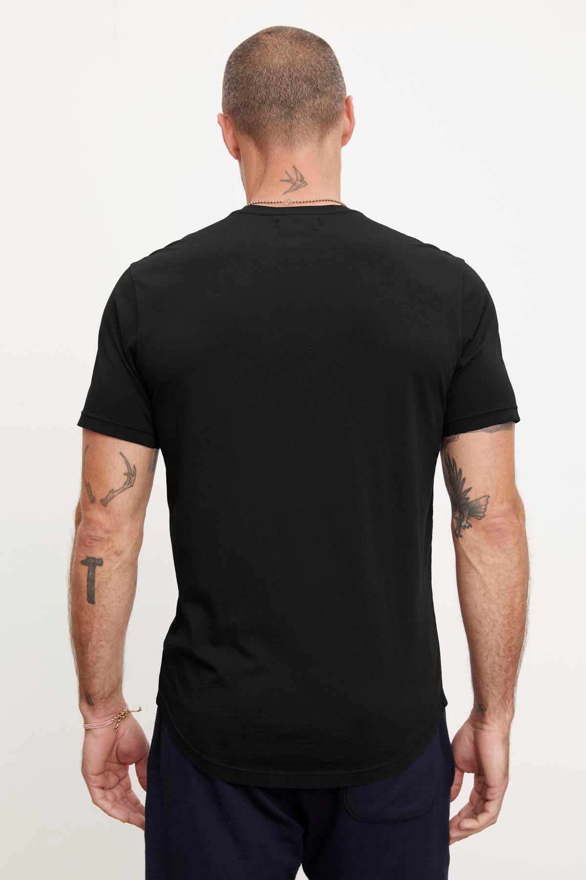   A man with a shaved head is standing with his back facing the camera, wearing a black FULTON HENLEY made of lightweight cotton jersey by Velvet by Graham & Spencer. He has several tattoos on both arms. 