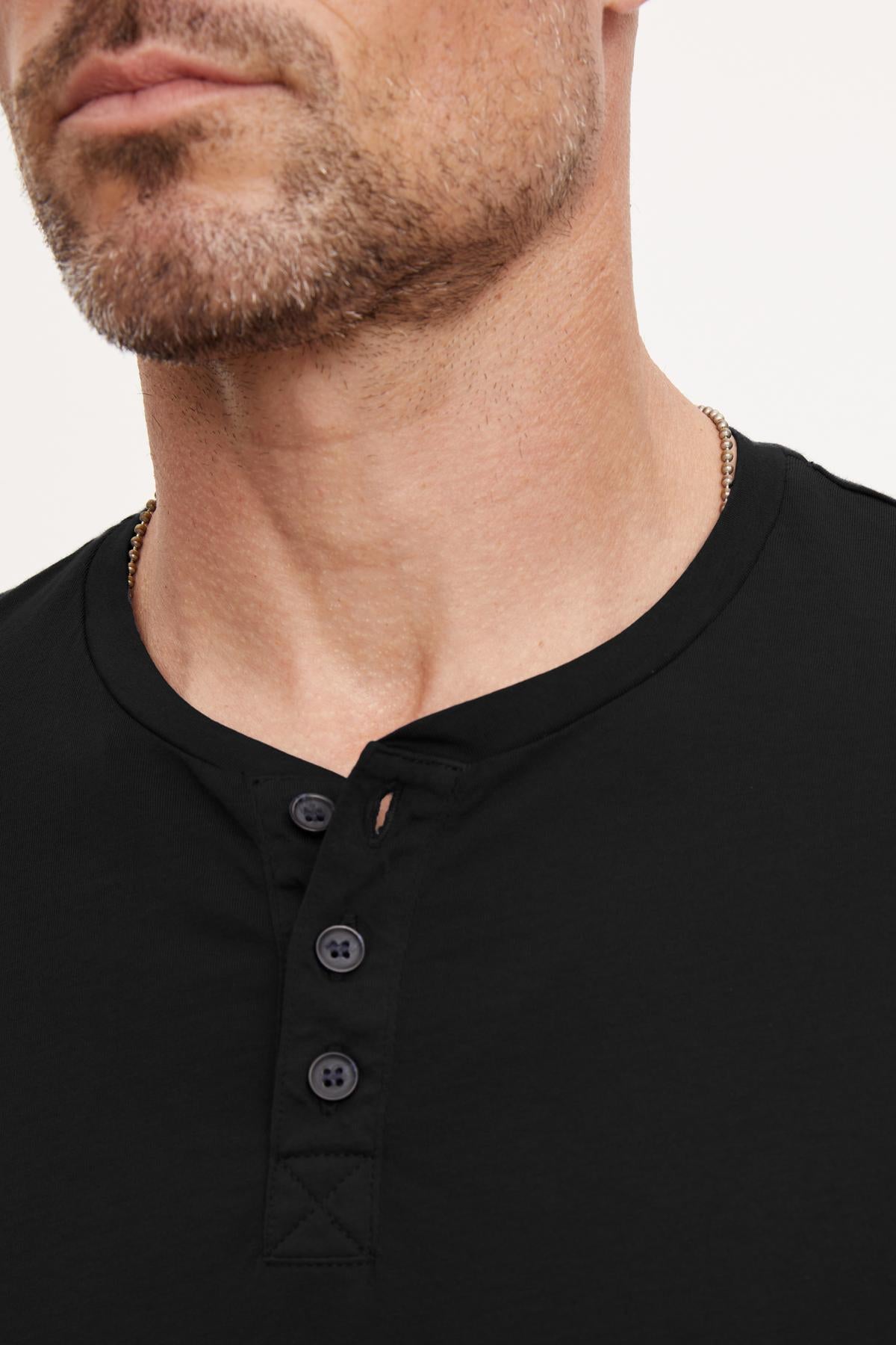   Close-up of a man with a trimmed beard wearing the FULTON HENLEY by Velvet by Graham & Spencer. The shirt features 3-buttons and a round neckline. His face is partially shown, focusing on the lightweight cotton jersey fabric and the curved hemline details. 