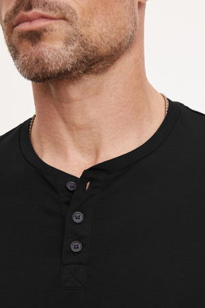 Close-up of a man with a trimmed beard wearing the FULTON HENLEY by Velvet by Graham & Spencer. The shirt features 3-buttons and a round neckline. His face is partially shown, focusing on the lightweight cotton jersey fabric and the curved hemline details.