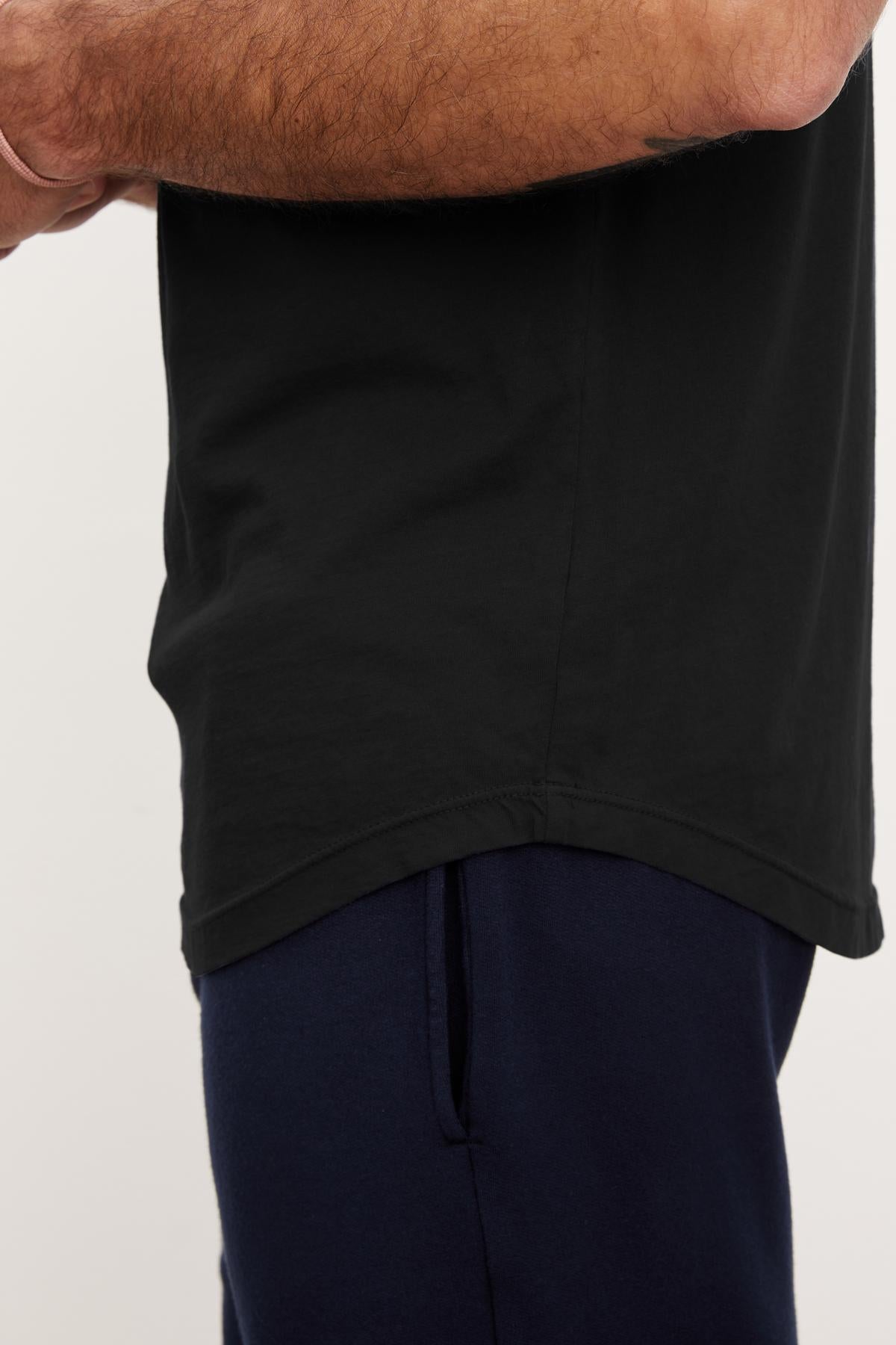   A person in a black FULTON HENLEY by Velvet by Graham & Spencer and navy pants, with their arms crossed, is partially visible from the shoulder to the waist against a plain background. 