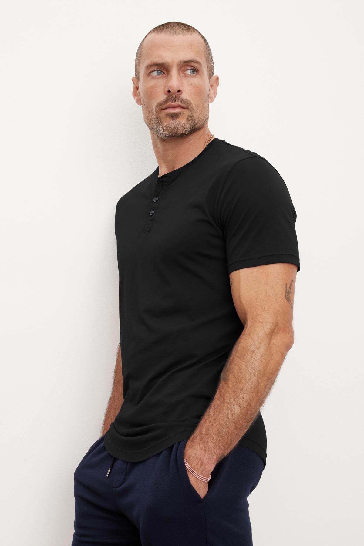   A man with a shaved head and short beard wears the FULTON HENLEY, a black, short-sleeved tee made of lightweight cotton jersey by Velvet by Graham & Spencer, paired with dark pants. He stands against a plain off-white background, looking to his left with hands in pockets as the curved hemline adds subtle sophistication. 