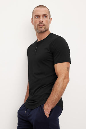 A man with a shaved head and short beard wears the FULTON HENLEY, a black, short-sleeved tee made of lightweight cotton jersey by Velvet by Graham & Spencer, paired with dark pants. He stands against a plain off-white background, looking to his left with hands in pockets as the curved hemline adds subtle sophistication.