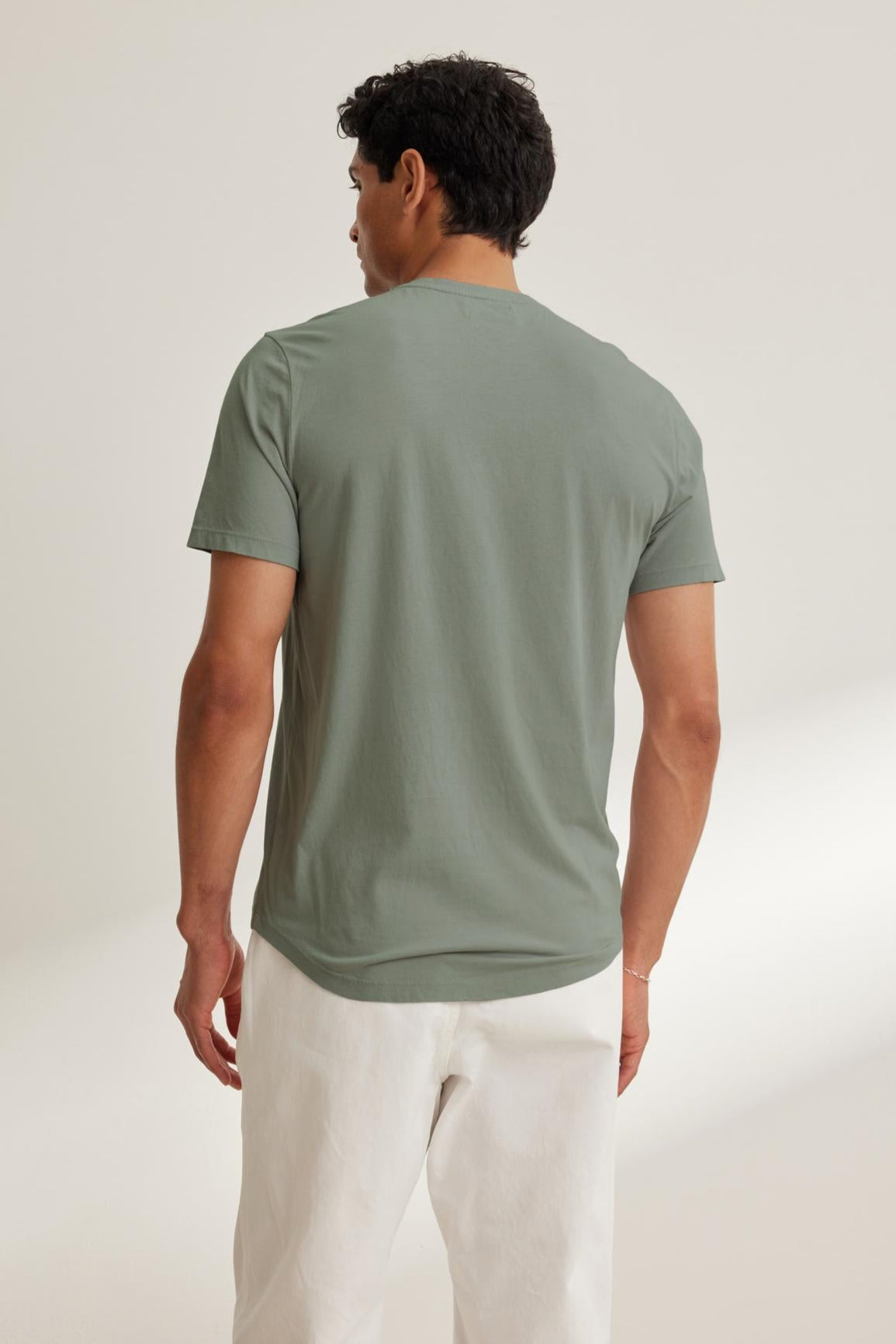   A man stands against a neutral background, wearing Velvet by Graham & Spencer's FULTON HENLEY in green, crafted from lightweight Pima jersey, paired with white pants. 