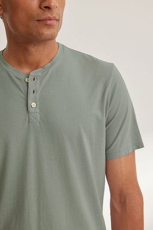 A person wearing the Velvet by Graham & Spencer FULTON HENLEY, a 3-button light green tee made of soft Pima jersey, is shown from the chest up, standing sideways.