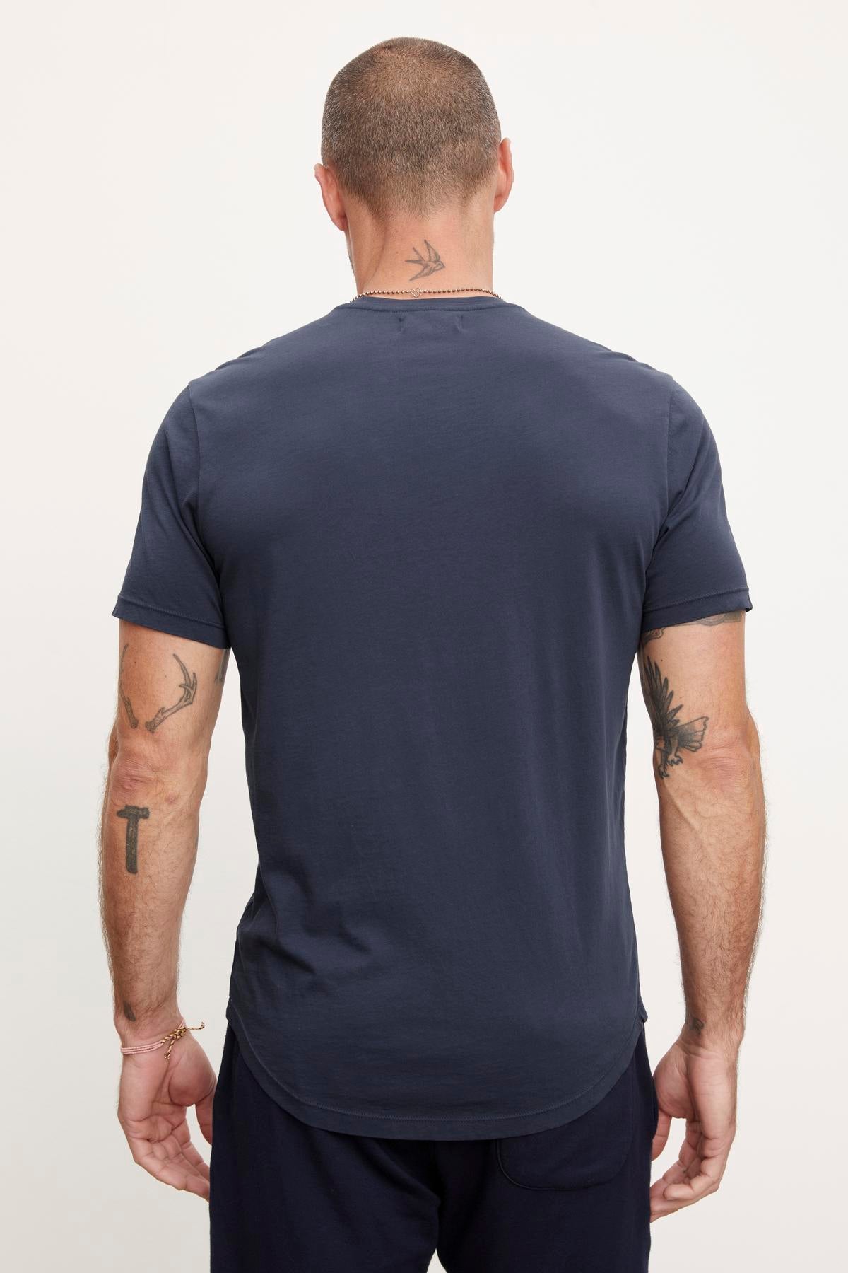 A man seen from behind wearing a dark blue Velvet by Graham & Spencer Fulton Henley tee, displaying multiple tattoos on his arms and a neck tattoo.-36753531109569