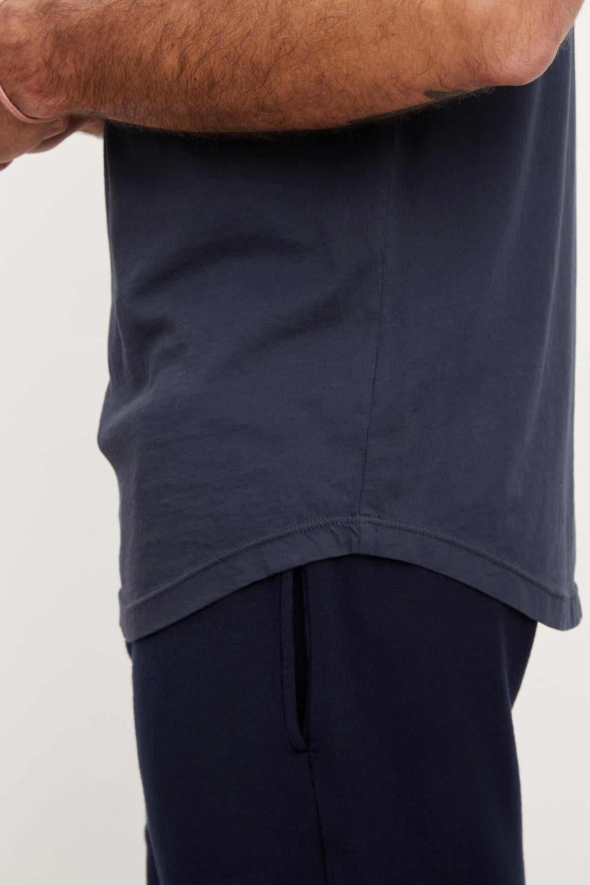   Close-up of a person holding up the hem of a plain navy blue t-shirt to reveal navy sweatpants, showcasing its curved hemline of the Fulton Henley by Velvet by Graham & Spencer. 