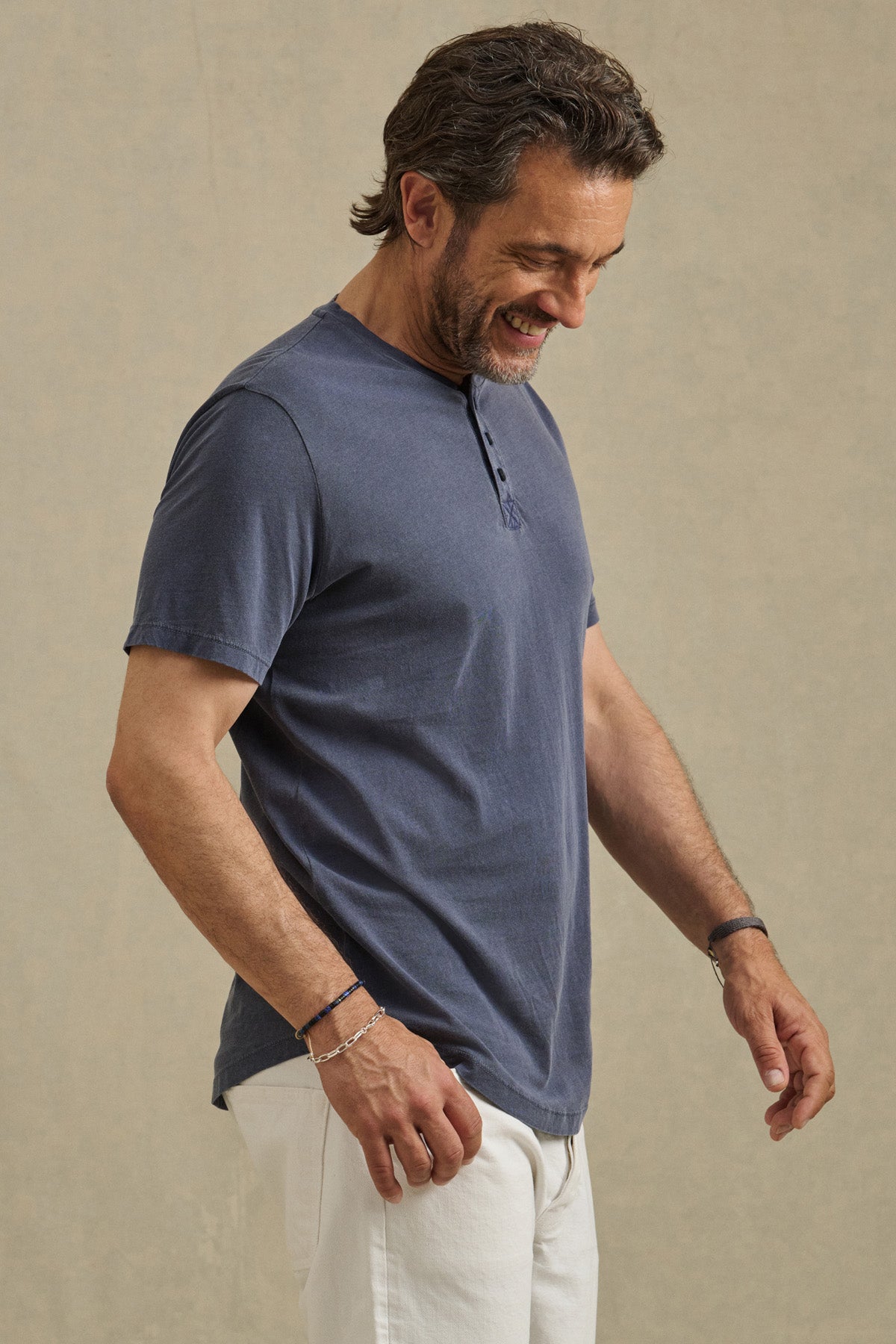 A man wearing the Velvet by Graham & Spencer FULTON HENLEY, a blue Pima jersey tee with a curved hemline, and white pants stands against a neutral background, looking down and smiling.-41626555580609