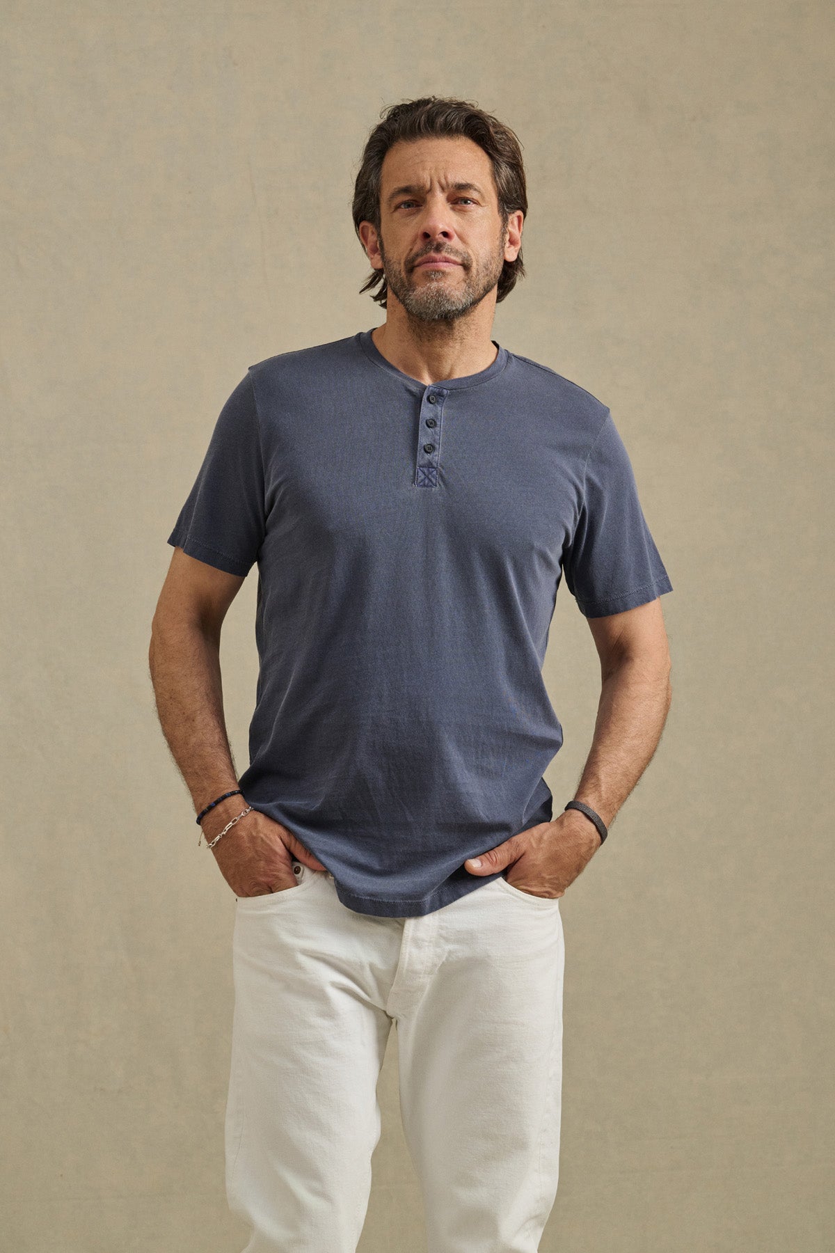 A man with dark hair and a beard stands against a neutral background, wearing the Velvet by Graham & Spencer FULTON HENLEY—a short-sleeved blue tee with a curved hemline—and white pants, his hands in his pockets.