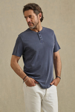 A man with a beard wears the FULTON HENLEY by Velvet by Graham & Spencer, a short-sleeved blue tee with a curved hemline, paired with white pants. He stands against a neutral background, one hand in his pocket, looking to the side.