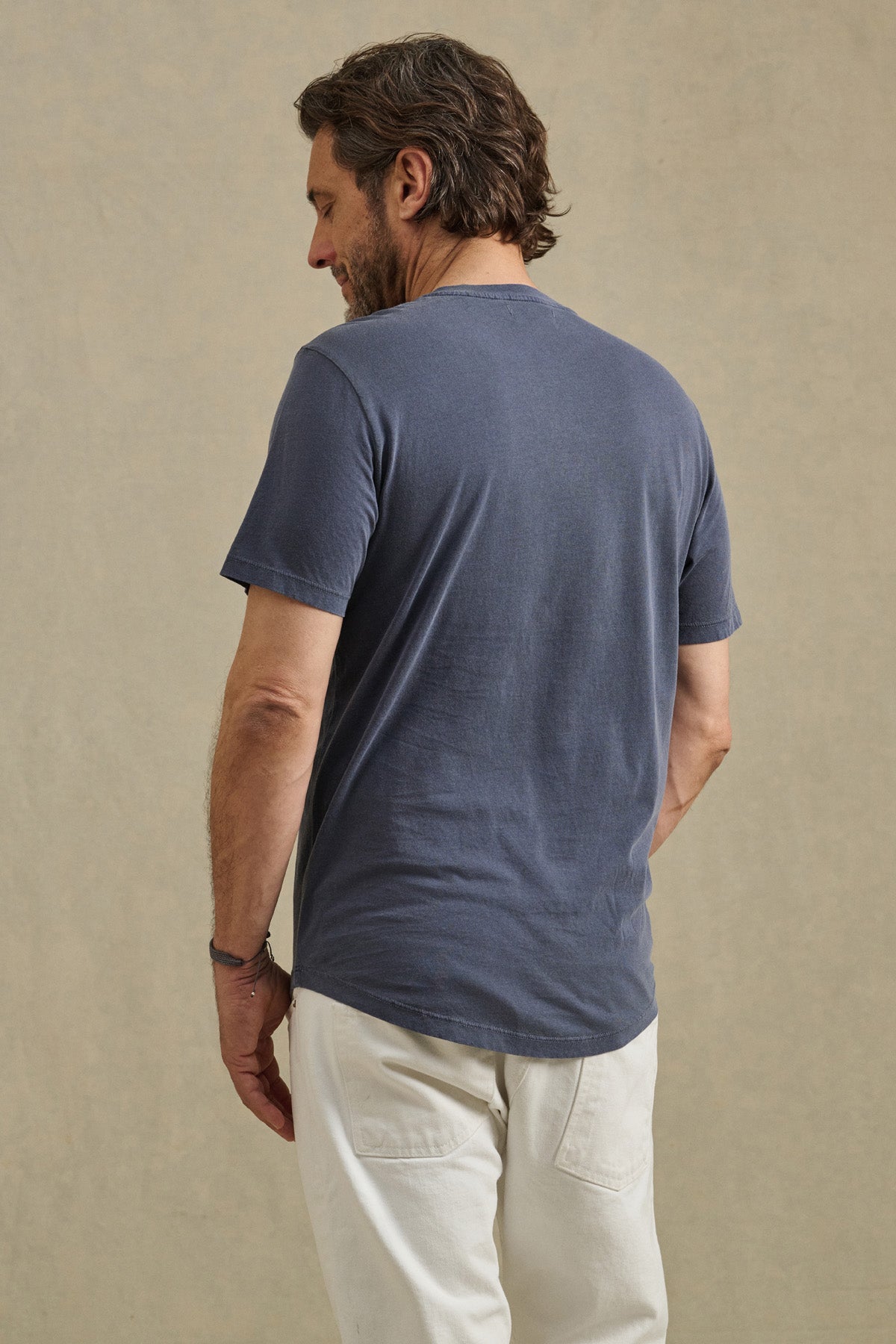 A man faces away, wearing the Velvet by Graham & Spencer FULTON HENLEY short-sleeve blue jersey tee and white pants against a neutral background.