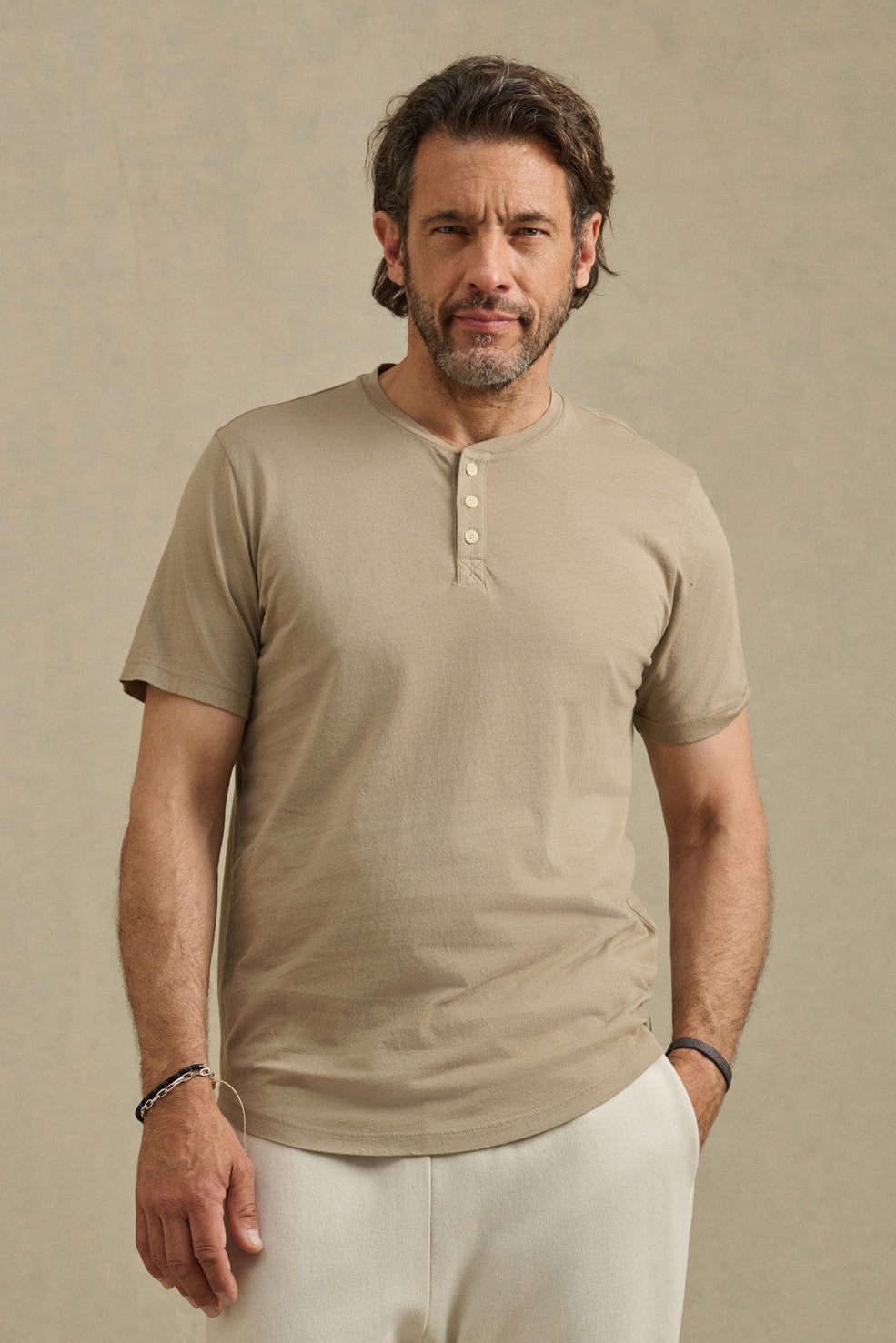 A man with medium-length hair and a beard wears the beige FULTON HENLEY by Velvet by Graham & Spencer and cream pants, standing against a plain neutral background.