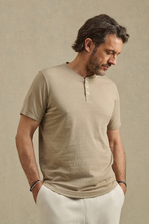 A man with medium-length hair and a beard wears Velvet by Graham & Spencer's FULTON HENLEY in beige Pima jersey, featuring a curved hem, paired with light pants, standing with hands in pockets against a neutral background.