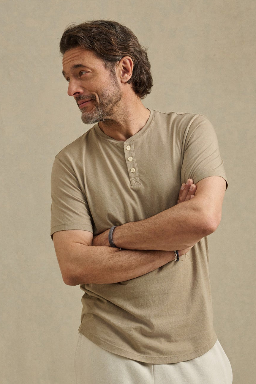 A man with wavy brown hair and a beard stands with arms crossed, looking to the side, wearing the FULTON HENLEY by Velvet by Graham & Spencer in beige and light pants against a plain background.