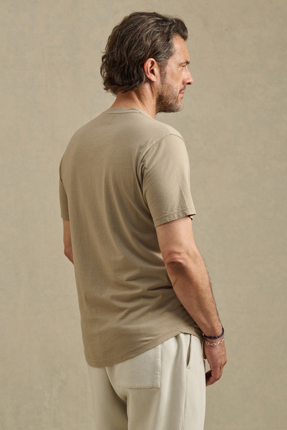 A man with short brown hair and a beard, wearing the FULTON HENLEY by Velvet by Graham & Spencer and light pants, stands in profile against a plain beige background.