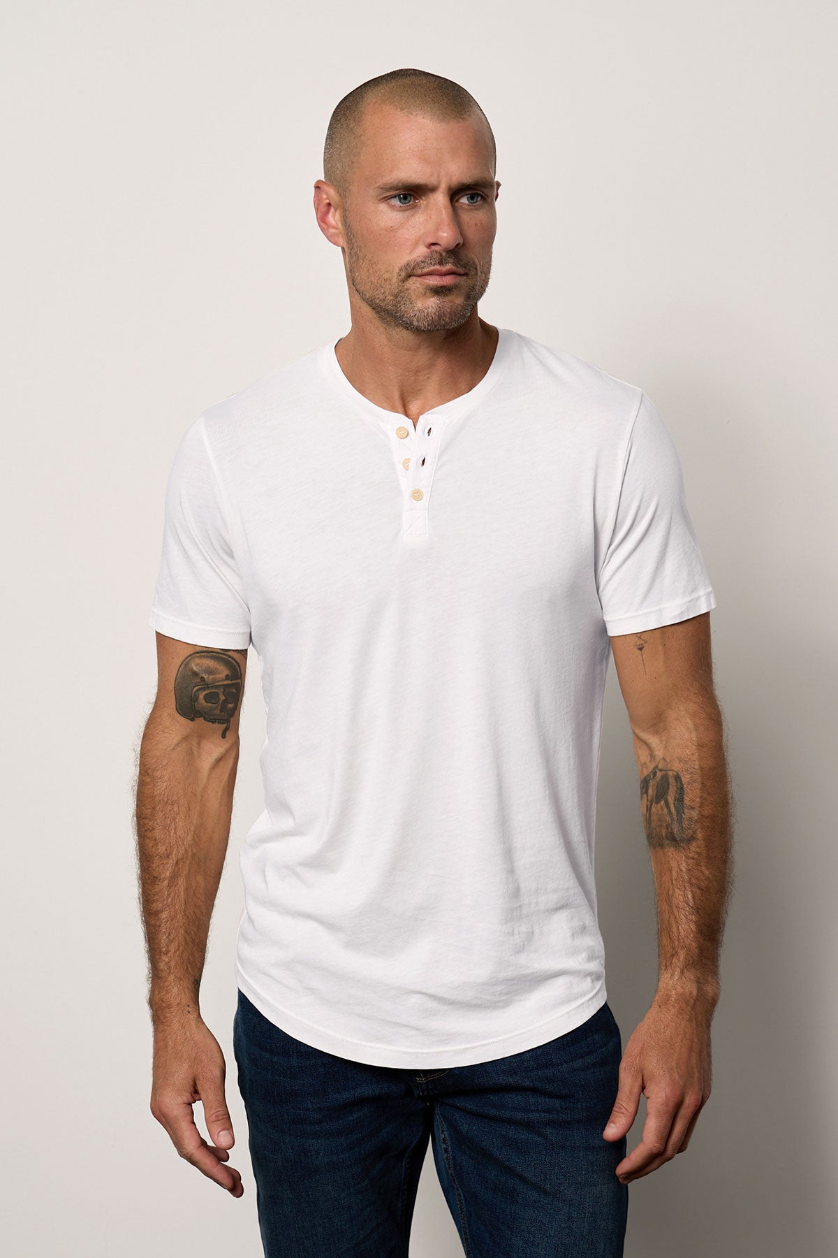   A man with a shaved head and tattoos on his arms, wearing a white Velvet by Graham & Spencer FULTON HENLEY tee, stands facing the camera with a neutral expression. 