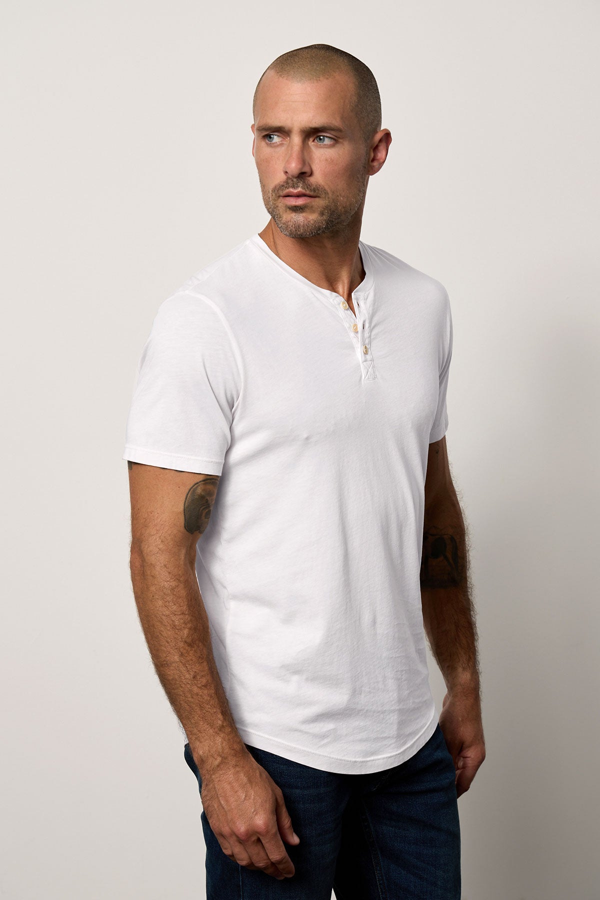   A man with a shaved head, wearing a white Velvet by Graham & Spencer Fulton Henley and dark jeans, stands against a plain background, looking to his left. 