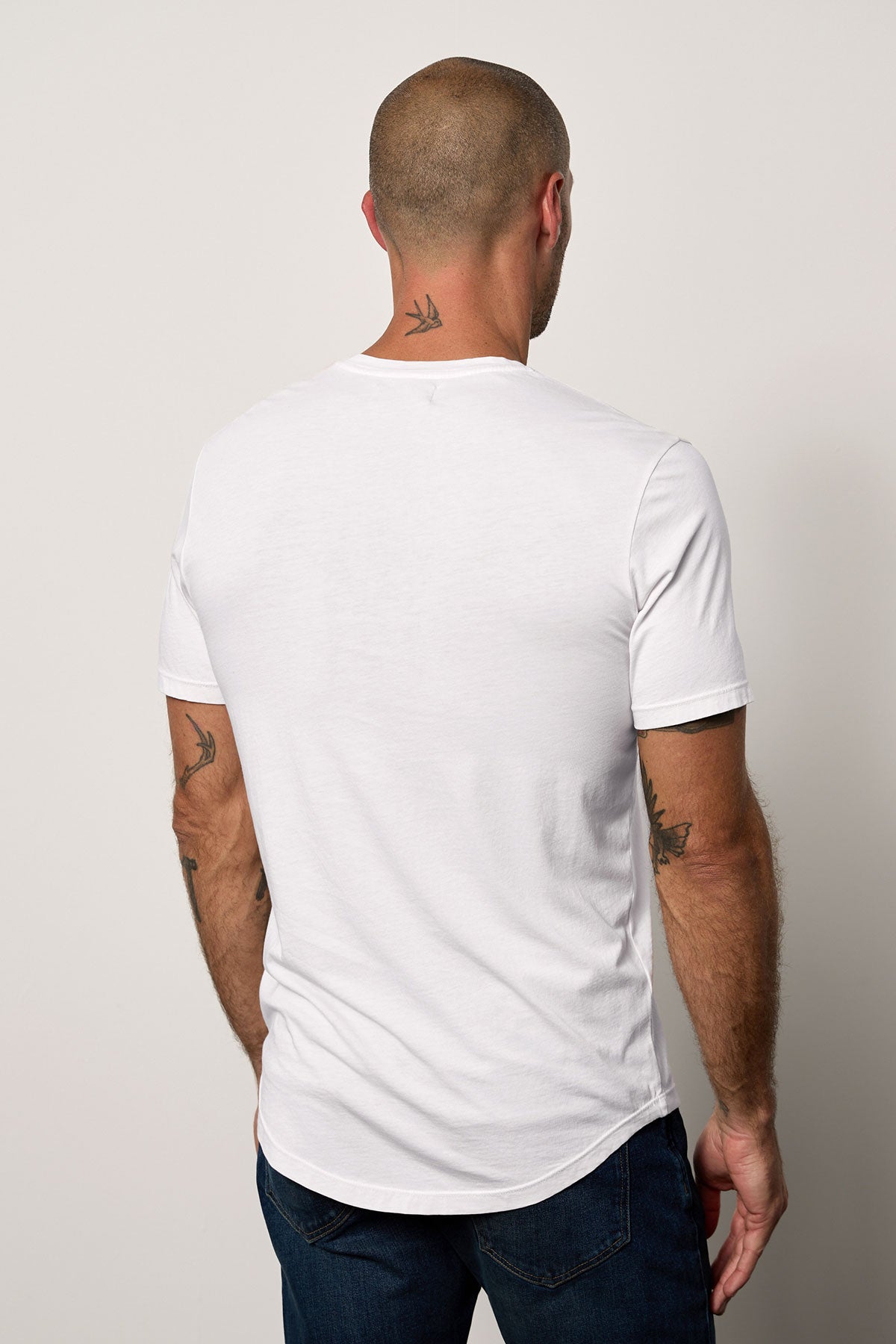 Man standing with his back to the camera, wearing a white Velvet by Graham & Spencer Fulton henley tee with a curved hemline, revealing tattoos on his arms and neck.-36640141181121