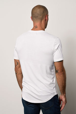 Man standing with his back to the camera, wearing a white Velvet by Graham & Spencer Fulton henley tee with a curved hemline, revealing tattoos on his arms and neck.