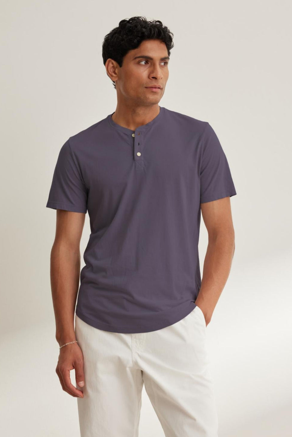   A man models the stylish curved hemline of the FULTON HENLEY by Velvet by Graham & Spencer, crafted from lightweight Pima jersey in dark gray, as he stands against a plain background with white pants. 