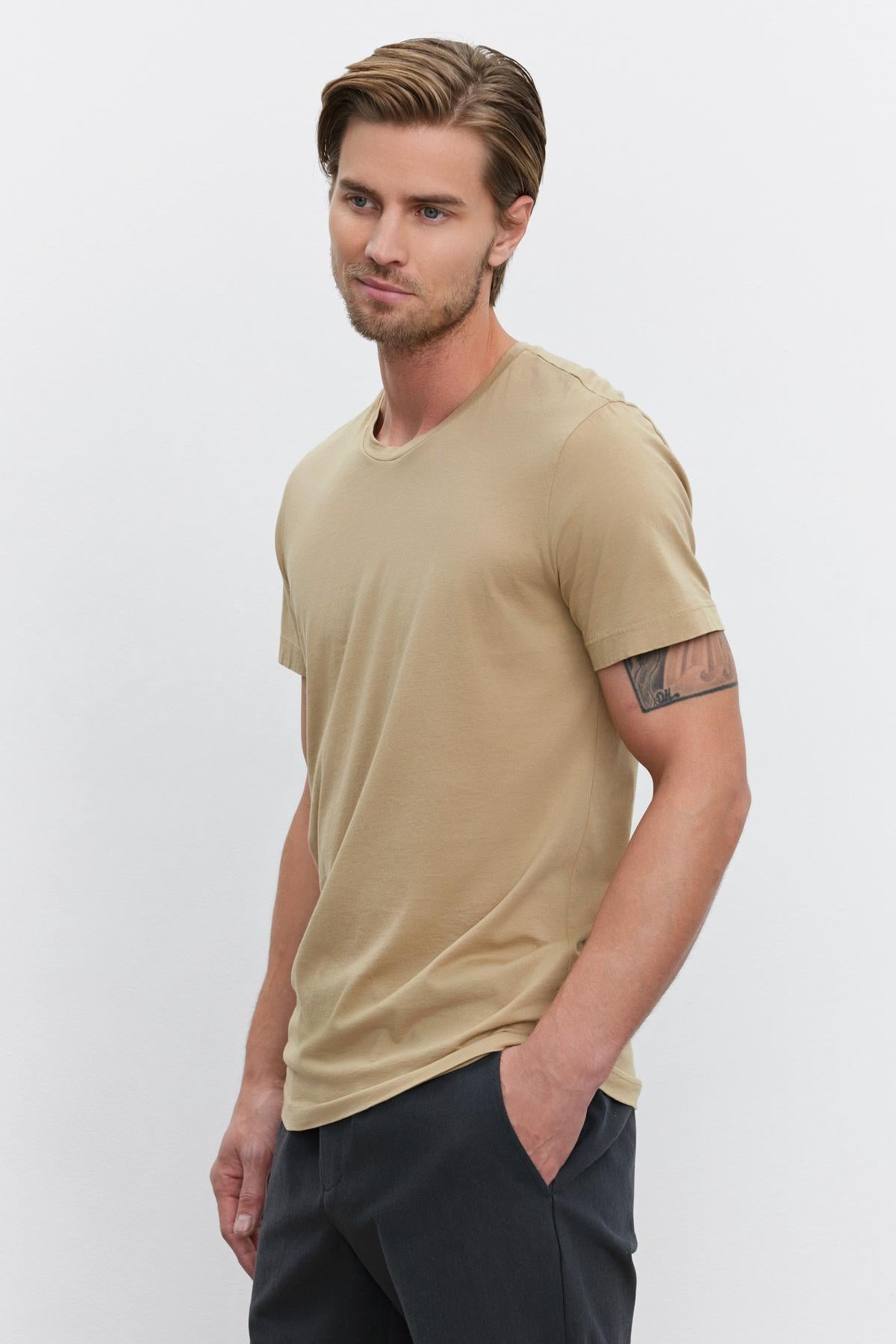   A man with short, styled hair and a tattoo on his left arm wearing a HOWARD TEE from Velvet by Graham & Spencer made of lightweight cotton knit and dark pants, standing with his hands in his pockets. 