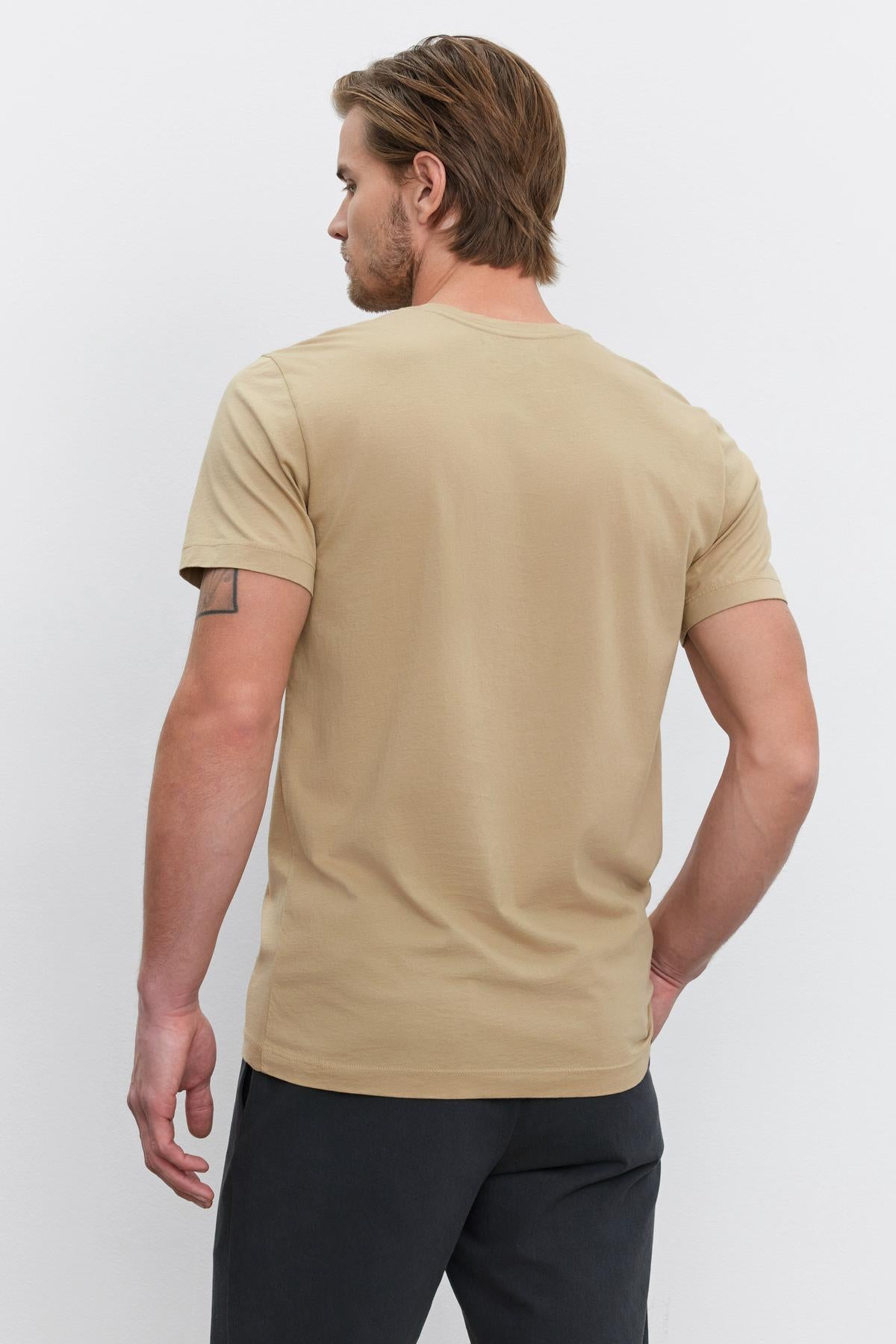   Back view of a man with brown hair wearing a beige HOWARD TEE by Velvet by Graham & Spencer made from lightweight cotton knit and dark pants, standing against a light background, giving off a vintage feel. 