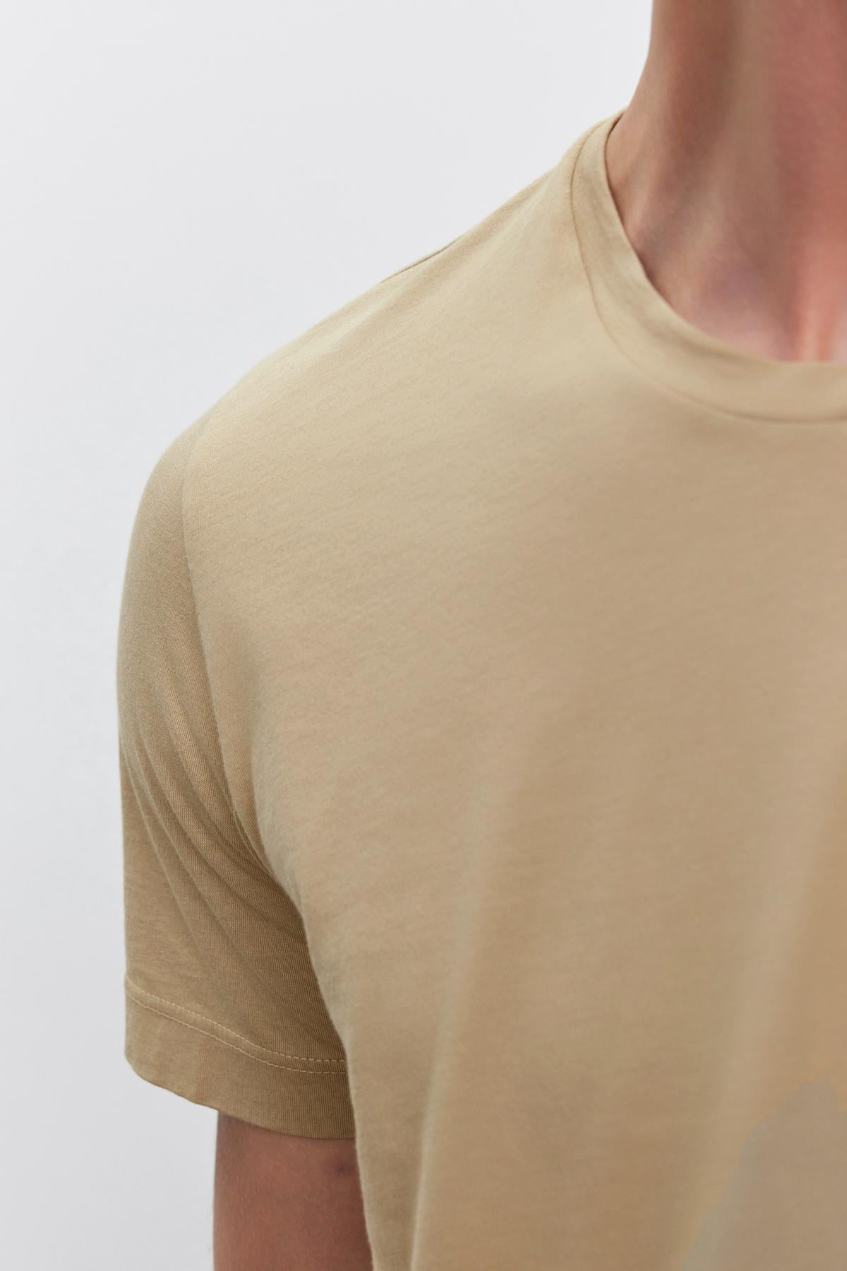   Close-up of a person wearing a beige HOWARD TEE made from lightweight cotton knit by Velvet by Graham & Spencer, showing the shoulder, arm, and part of the neck against a plain white background. 