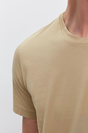 Close-up of a person wearing a beige HOWARD TEE made from lightweight cotton knit by Velvet by Graham & Spencer, showing the shoulder, arm, and part of the neck against a plain white background.