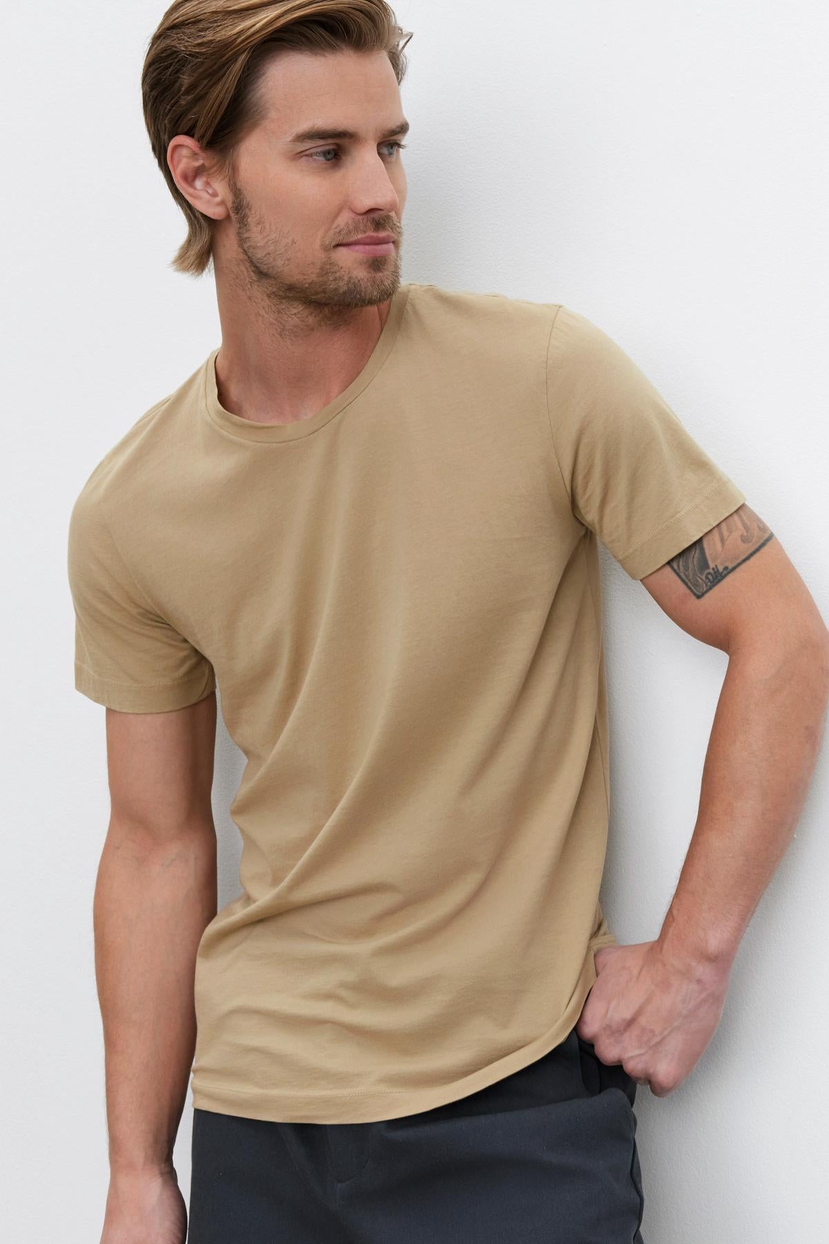   A person wearing a beige HOWARD TEE by Velvet by Graham & Spencer made of lightweight cotton knit and dark pants stands against a plain light-colored background, looking to the side. 