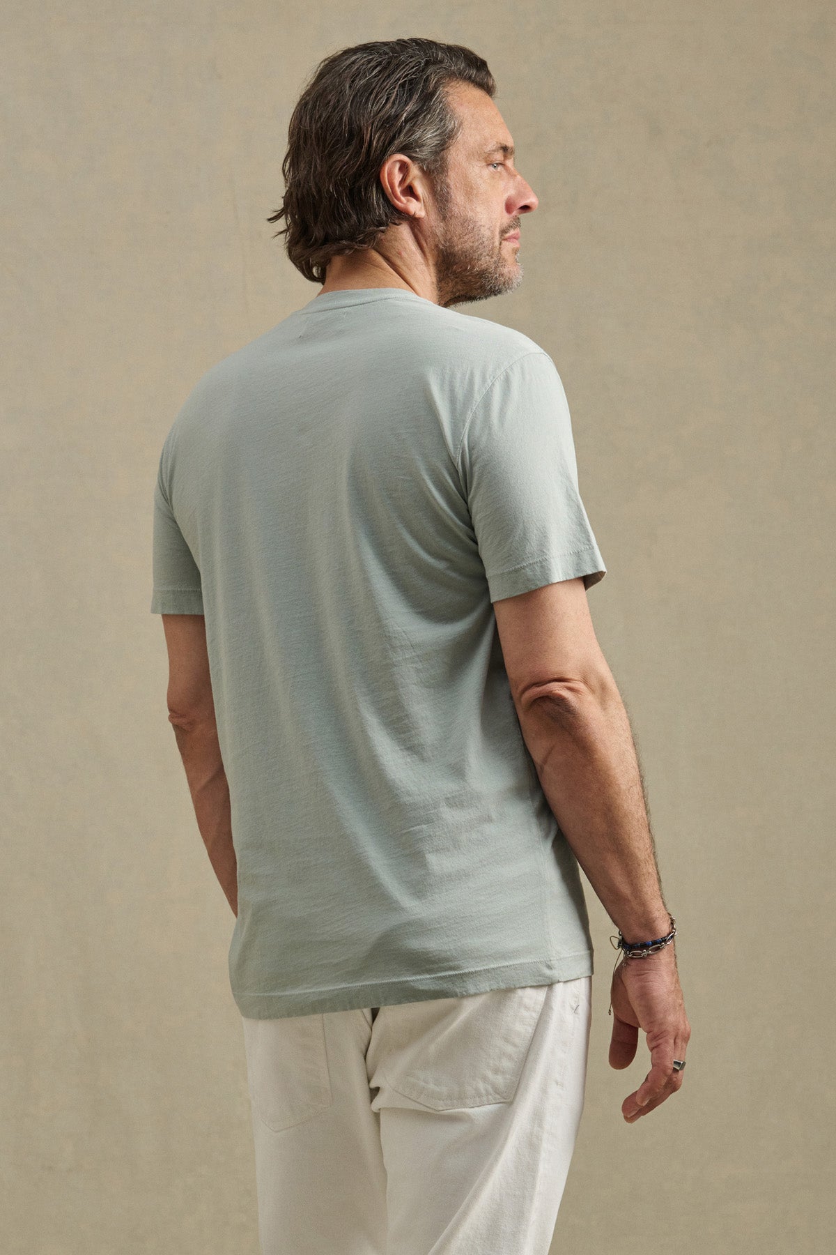 A man with short brown hair models the Velvet by Graham & Spencer HOWARD TEE, a light gray Pima jersey shirt, paired with white pants. He stands with his back to the camera against a neutral background, highlighting its perfect fit.