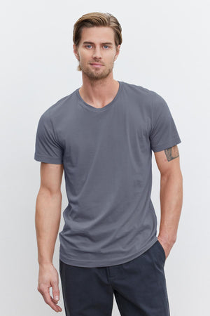 A person wearing a gray HOWARD TEE made of lightweight cotton knit from Velvet by Graham & Spencer and dark pants stands against a plain white background. One arm is resting by their side while the other is partially in their pocket, creating a relaxed yet perfect fit with a hint of vintage feel.