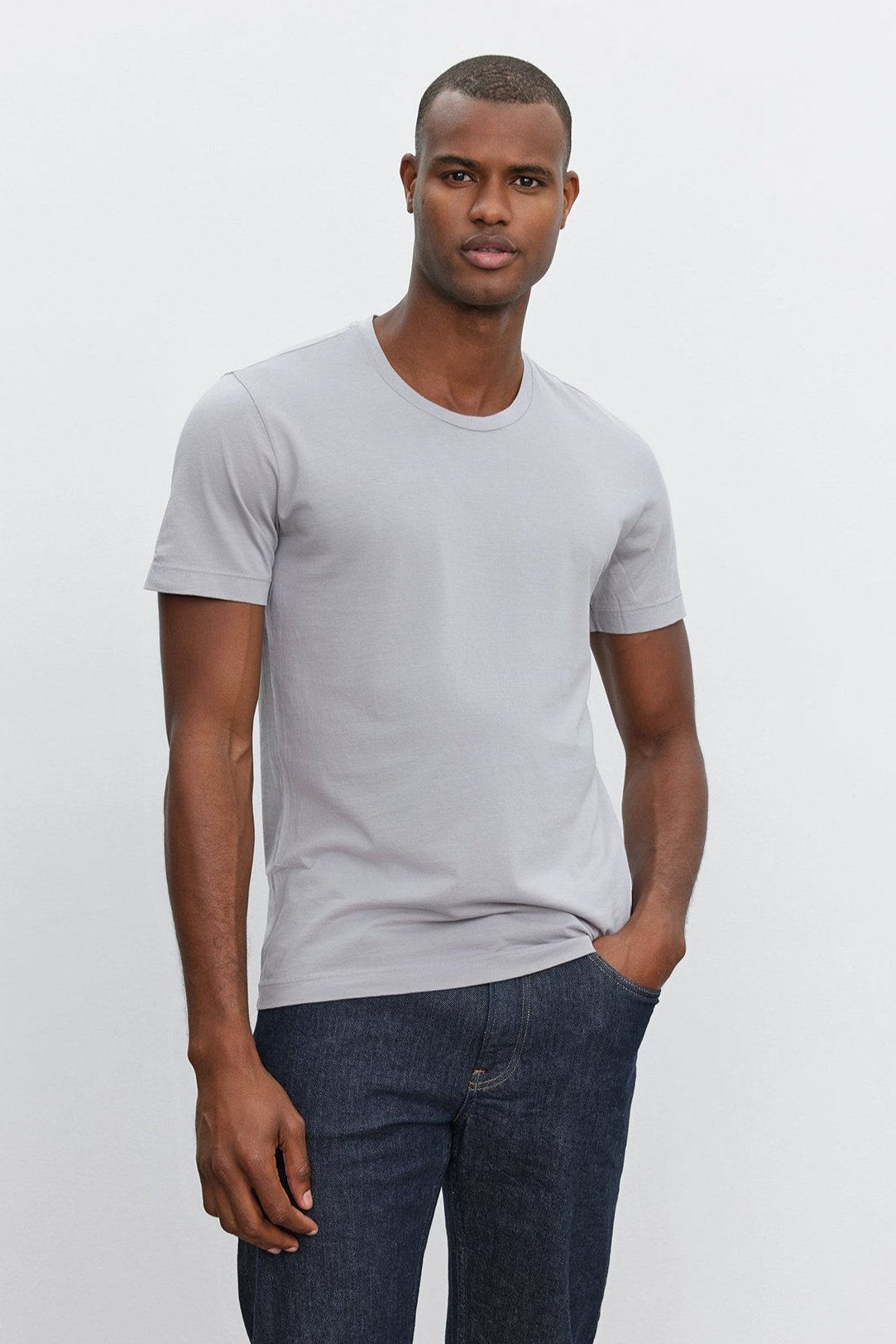   A person stands against a plain background, wearing a light gray HOWARD TEE from Velvet by Graham & Spencer made from lightweight cotton knit and dark blue jeans that boast the perfect fit. 