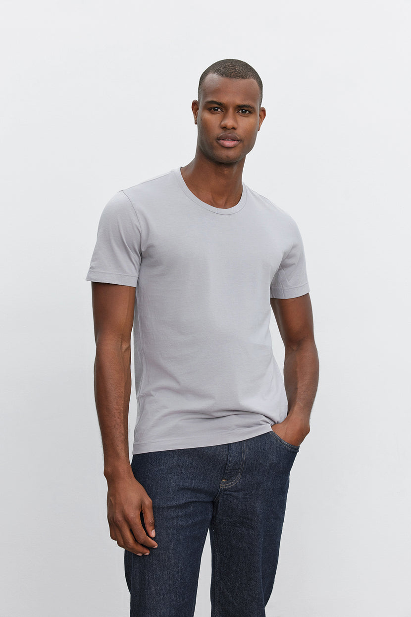 A person stands against a plain background, wearing a light gray HOWARD TEE from Velvet by Graham & Spencer made from lightweight cotton knit and dark blue jeans that boast the perfect fit.