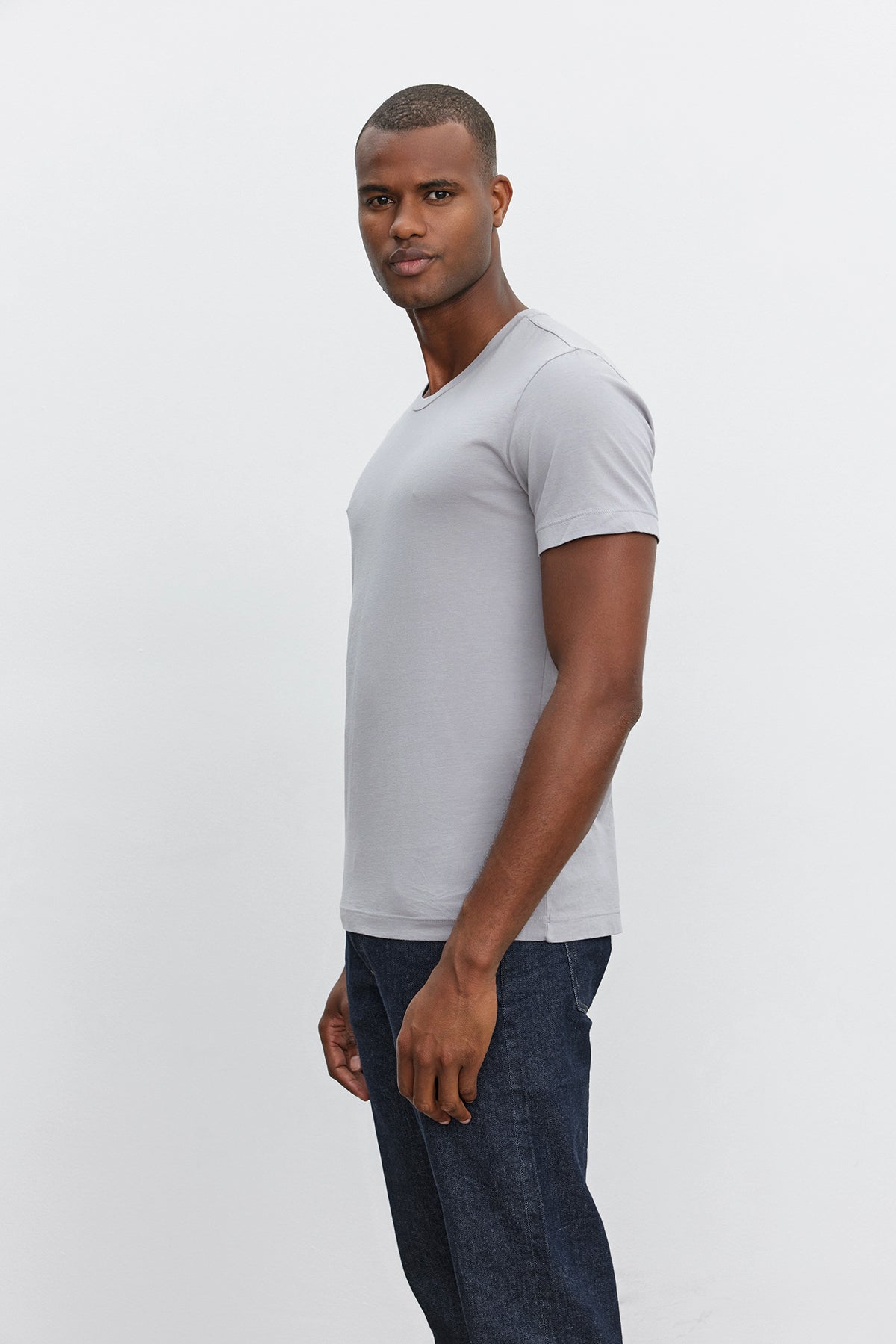 A man wearing a light gray HOWARD TEE by Velvet by Graham & Spencer in lightweight cotton knit and dark blue jeans with a vintage feel stands against a plain white background, looking at the camera.-37386165289153