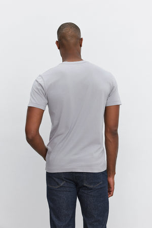 Back view of a person wearing the HOWARD TEE by Velvet by Graham & Spencer, a lightweight cotton knit light gray short-sleeve t-shirt, and dark blue jeans against a plain white background.