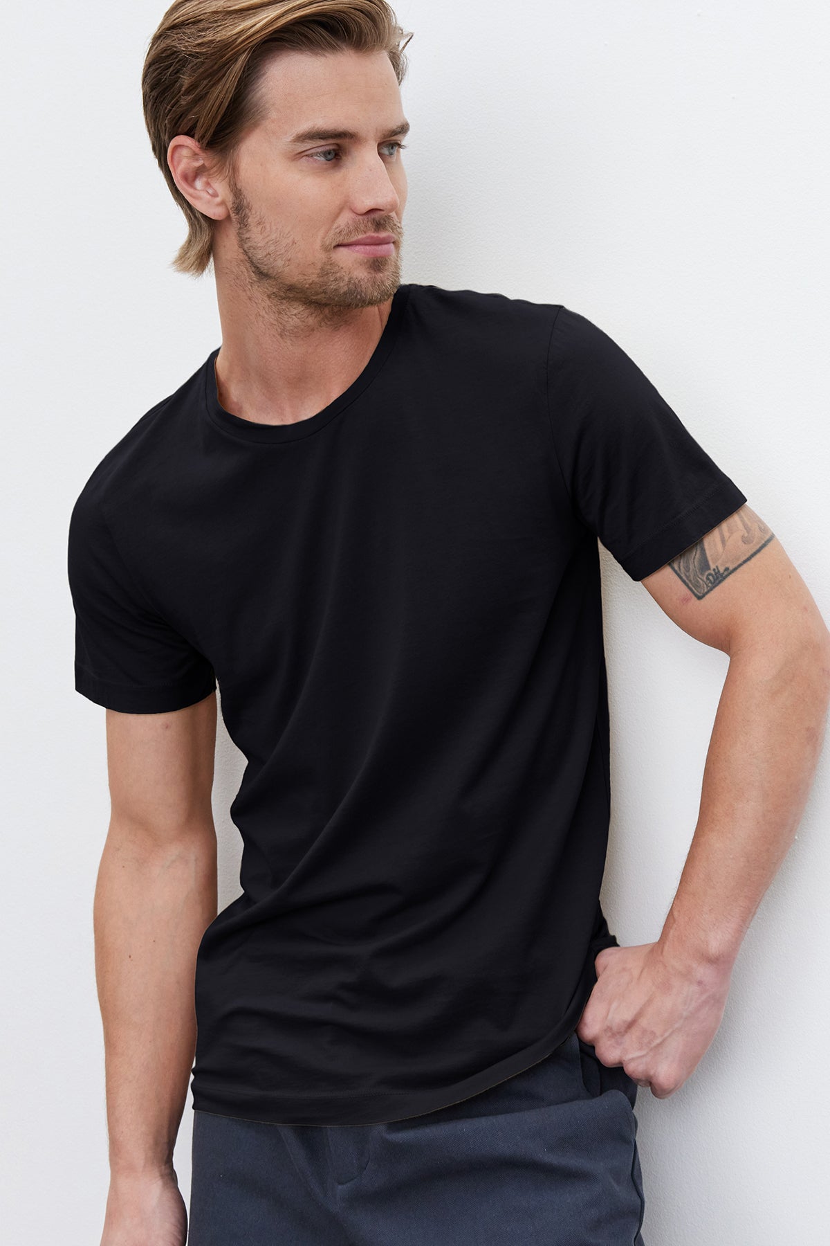 A man in a black Velvet by Graham & Spencer HOWARD WHISPER CLASSIC CREW NECK TEE leaning against a wall.-36162036891841