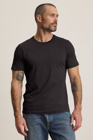 A bearded man in blue jeans and a black HOWARD TEE from Velvet by Graham & Spencer, made of lightweight Pima jersey, stands with his hands at his sides, looking to the right. The perfect fit complements his vintage style.