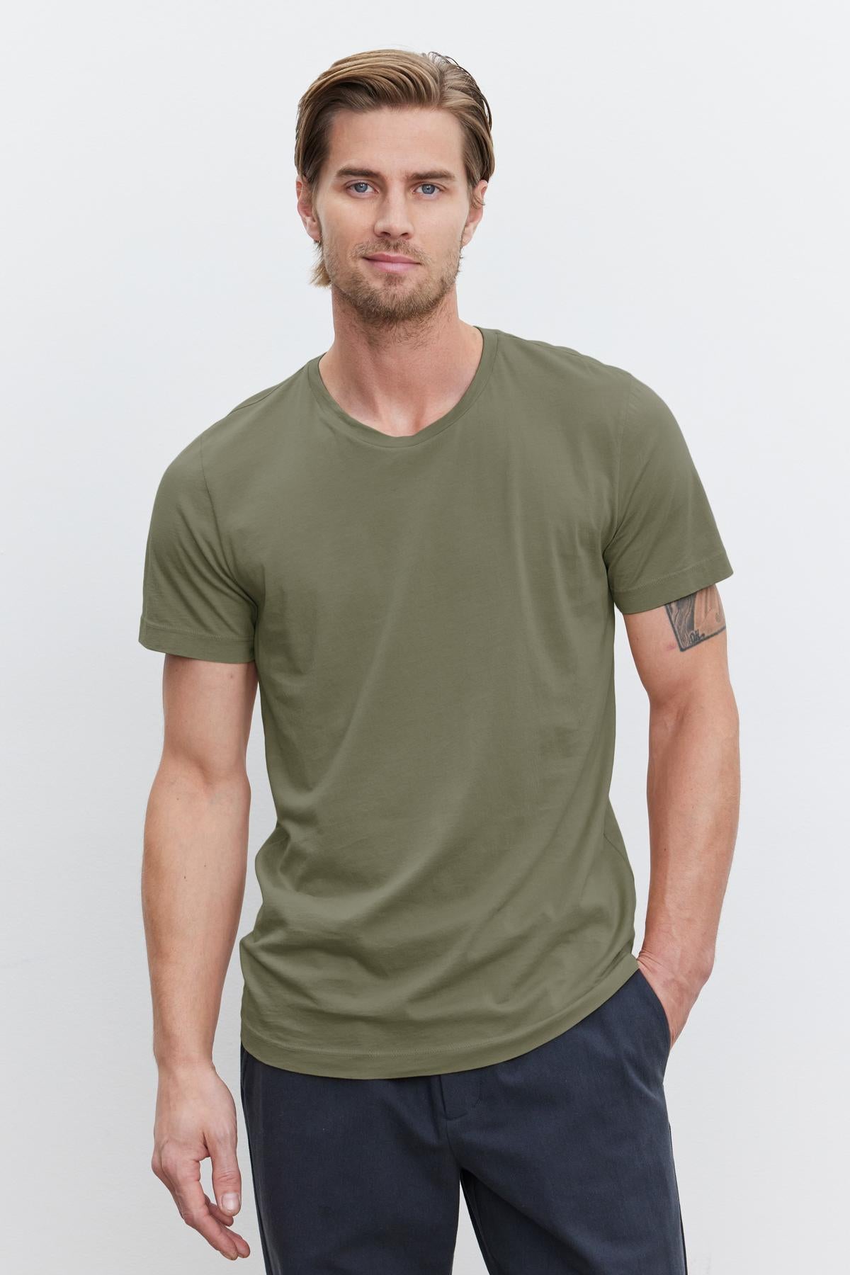 A man with light brown hair and a beard is standing against a plain background, wearing a green Velvet by Graham & Spencer HOWARD TEE in lightweight cotton knit and dark pants.-37386103455937