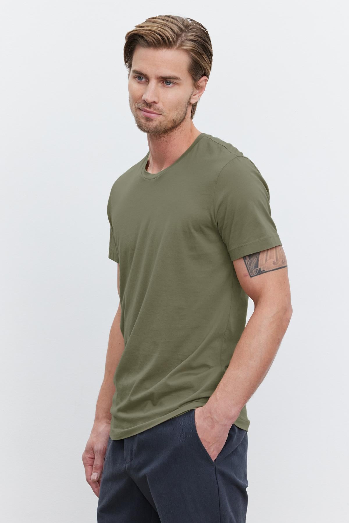 A young man with light brown hair wearing an olive green HOWARD TEE from Velvet by Graham & Spencer made from lightweight cotton knit and dark pants stands with his hands in his pockets against a plain white background, giving off a perfect fit.-37386103423169