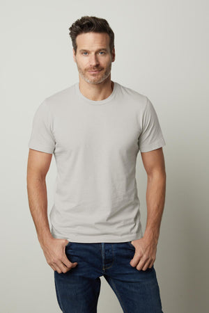 A man with short, dark hair and a beard wearing the light gray Velvet by Graham & Spencer HOWARD TEE and blue jeans stands with his hands in his pockets, exuding a vintage feel against a plain background.