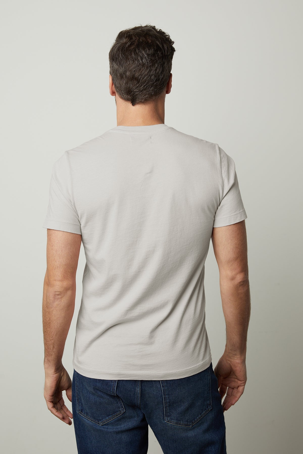   A man with short, dark hair stands with his back to the camera, wearing a light gray Velvet by Graham & Spencer HOWARD TEE made from lightweight cotton knit and dark blue jeans, offering a perfect fit. 