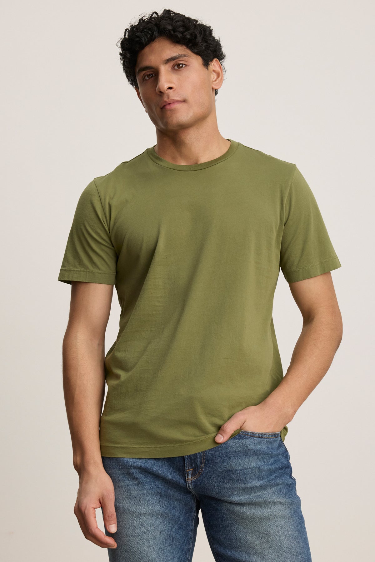 A man wears the HOWARD TEE by Velvet by Graham & Spencer, a plain olive green lightweight Pima jersey t-shirt, with blue jeans, standing against a neutral background with one hand in his pocket.-39446027763905